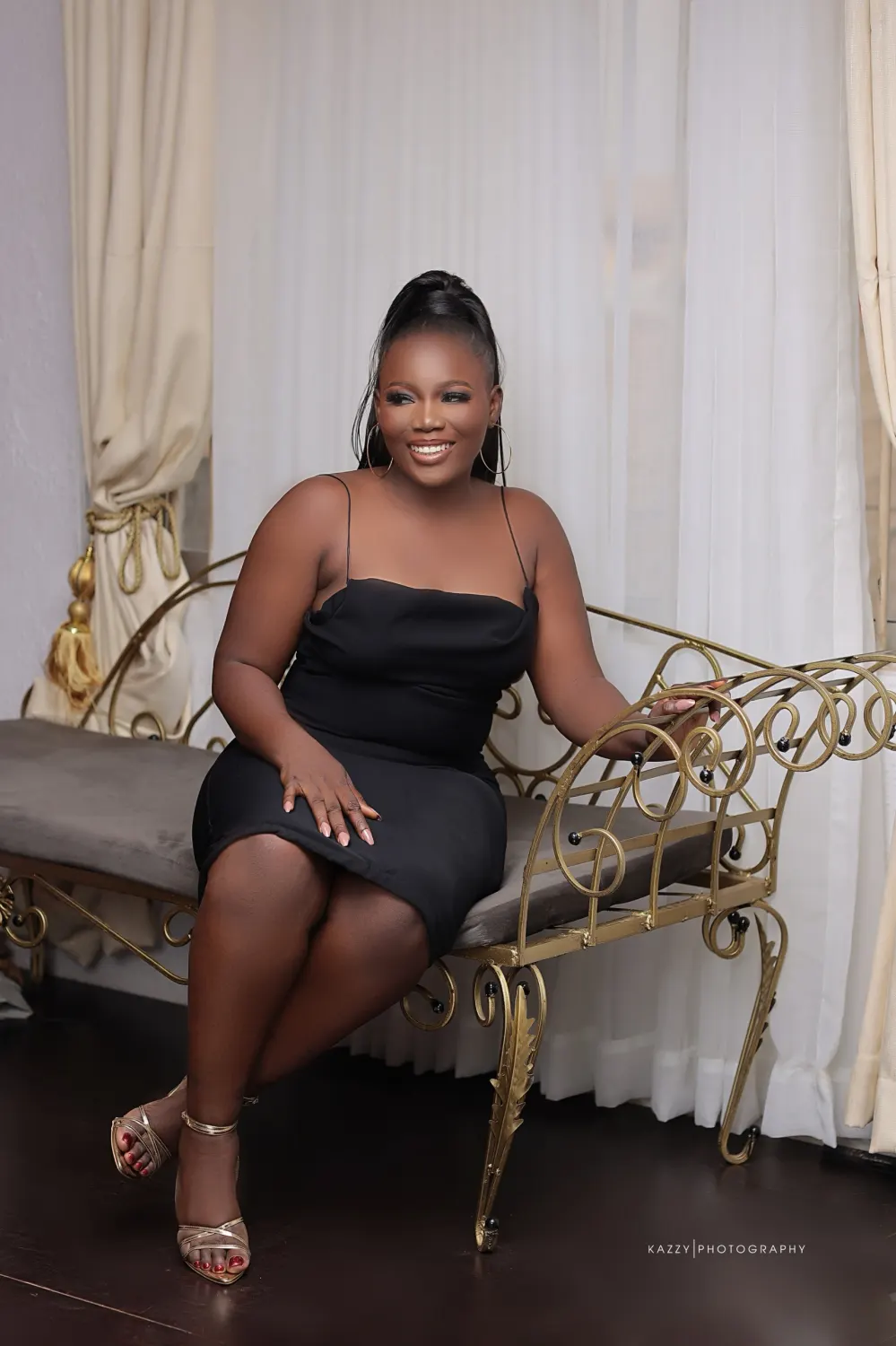 Adediwura laughing, seated on a gold bench in front of cream curtains