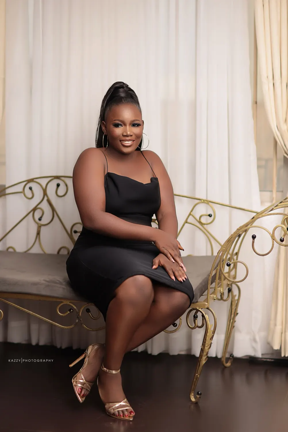 Adediwura in a black dress and gold heels, seated on a gold wrought-iron bench, smiling